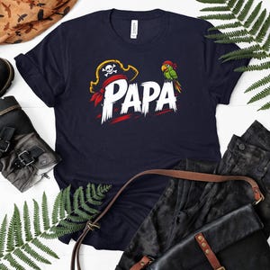 May include: Navy blue t-shirt with a pirate-themed design. The shirt features the word "PAPA" in white, with a pirate hat and a parrot graphic. The design includes red accents.