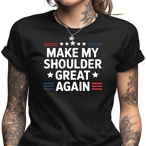 May include: Black t-shirt with the white text "MAKE MY SHOULDER GREAT AGAIN" in a bold, sans-serif font. The text is framed by red and blue horizontal lines and stars, creating a patriotic design.