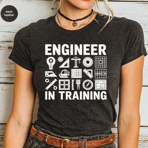 May include: Dark heather gray t-shirt with the words "ENGINEER IN TRAINING" in white block letters. The shirt features engineering-related icons. The shirt has a crew neck and short sleeves.