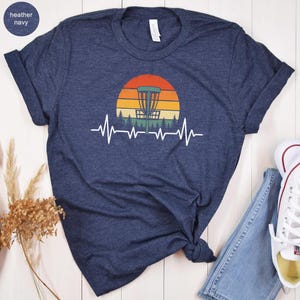 May include: Heather navy t-shirt featuring a disc golf basket design set against a sunset backdrop. A white heartbeat line graphic runs below the basket. The shirt has rolled-up sleeves and is paired with jeans and sneakers.
