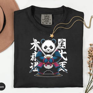 May include: Black t-shirt featuring a panda warrior design with crossed swords and Japanese text. The panda is wearing blue and red armor. A gold necklace with a star pendant is draped on the shirt.