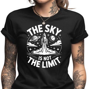 May include: Black t-shirt with a white graphic of a rocket launching into space, surrounded by stars, planets, and clouds. The text "THE SKY IS NOT THE LIMIT" is printed in a circular design.