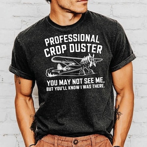 May include: Dark gray t-shirt with the text "PROFESSIONAL CROP DUSTER" above an illustration of a small airplane. Below the plane, the text reads "YOU MAY NOT SEE ME, BUT YOU'LL KNOW I WAS THERE."