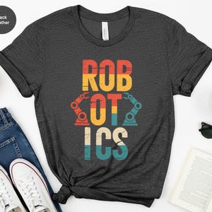 May include: Black heather t-shirt with the word "ROBOTICS" in a retro, colorful design. The word "ROBOTICS" is in a bold, blocky font with a robot arm graphic. The shirt is paired with jeans and white sneakers.