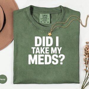 May include: Olive green t-shirt with the text "DID I TAKE MY MEDS?" in white block letters. A gold necklace with a star pendant rests on the shirt. A brown hat and sprigs of dried flowers are in the background.
