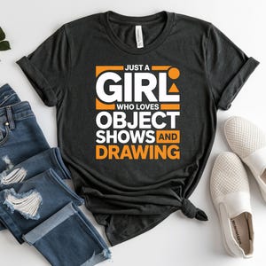 May include: Dark gray t-shirt with the text "JUST A GIRL WHO LOVES OBJECT SHOWS AND DRAWING" in white and orange. The shirt is paired with blue jeans and white slip-on shoes.