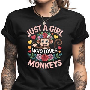 May include: Black t-shirt with a colorful graphic. The graphic features a smiling cartoon monkey surrounded by flowers and hearts, with the text "JUST A GIRL WHO LOVES MONKEYS" in pink and white.