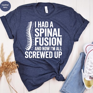 May include: Heather navy blue t-shirt with the text "I HAD A SPINAL FUSION AND NOW I'M ALL SCREWED UP" in white. A white spine graphic is on the left side of the text. The shirt is folded at the bottom.