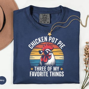 May include: Navy blue t-shirt with a graphic of a rooster wearing sunglasses, the text "CHICKEN POT PIE THREE OF MY FAVORITE THINGS" is printed on the front. The shirt is a Comfort Colors XL-TO-EG.