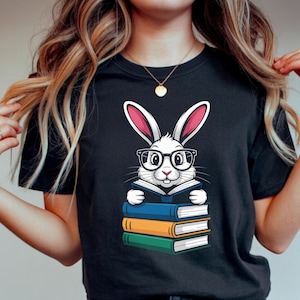 May include: Black t-shirt featuring a cartoon rabbit wearing glasses and reading a book. The rabbit is white with pink ears and is sitting on top of a stack of books in blue, orange, and green.