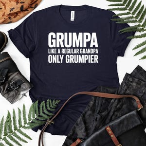 May include: Navy blue t-shirt with the text "GRUMPA LIKE A REGULAR GRANDPA ONLY GRUMPIER" in white block letters. The shirt is displayed with black pants, a leather bag, and a pair of boots.