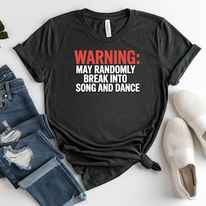 May include: Dark gray t-shirt with the text "WARNING: MAY RANDOMLY BREAK INTO SONG AND DANCE" in red and white. The shirt is paired with blue jeans and white slip-on shoes.