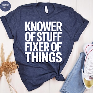May include: Heather navy blue t-shirt with the text "KNOWER OF STUFF FIXER OF THINGS" in white block letters. The shirt is styled with rolled-up sleeves and a knot at the waist. A pair of white sneakers and blue jeans are visible.