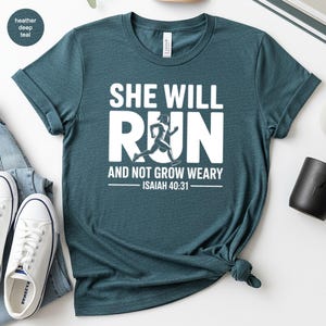 May include: Heather deep teal t-shirt with the text "SHE WILL RUN AND NOT GROW WEARY - ISAIAH 40:31" in white. The shirt features a graphic of a running figure. The shirt is made of a soft material and has short sleeves.