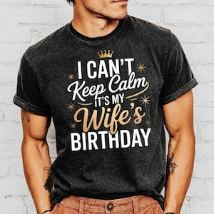 May include: Dark gray t-shirt with the text "I CAN'T Keep Calm IT'S MY Wife's BIRTHDAY" in white and gold lettering. The design includes a small gold crown and starburst accents. The shirt has rolled-up sleeves.