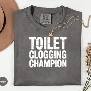 May include: A gray t-shirt with the text "TOILET CLOGGING CHAMPION" in white block letters. The shirt has a crew neck and a tag that says "Comfort Colors". A gold necklace with a star pendant is draped across the shirt.
