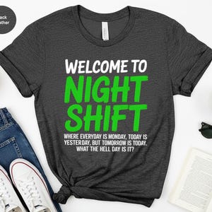 May include: Black heather t-shirt with the text "WELCOME TO NIGHT SHIFT" in white and green. The shirt also has the text "WHERE EVERYDAY IS MONDAY, TODAY IS YESTERDAY, BUT TOMORROW IS TODAY. WHAT THE HELL DAY IS IT?"