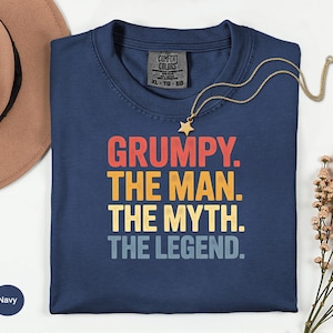 May include: Navy blue t-shirt with the text "GRUMPY. THE MAN. THE MYTH. THE LEGEND." in red, yellow, and blue lettering. A gold necklace with a star pendant rests on the shirt. A brown hat is visible in the background.