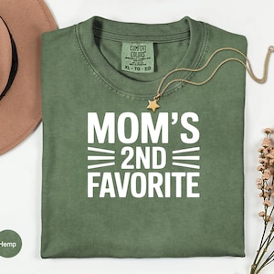 May include: Olive green t-shirt with the text "MOM'S 2ND FAVORITE" in white. A gold necklace with a star pendant rests on the shirt. A brown hat and a sprig of dried flowers are in the background.