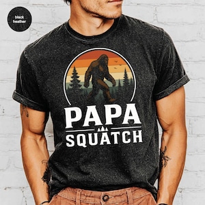 May include: Black heather t-shirt featuring a Bigfoot graphic inside a circular design with a sunset background and pine trees. The text "PAPA SQUATCH" is printed below the graphic in white block letters. The shirt has rolled-up sleeves.