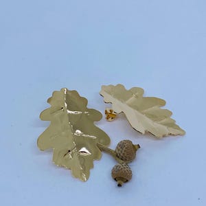 Può includere: Un paio di orecchini a forma di foglia di quercia dorati con due piccole ghiande.