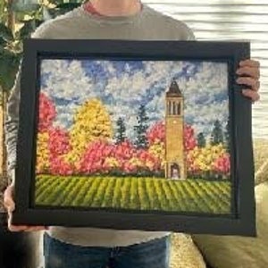 May include: A framed landscape painting featuring a tall, tan bell tower standing amidst a vibrant forest of yellow, orange, and pink autumn trees under a blue sky with white clouds, set above a field of green grass with textured brushstrokes.