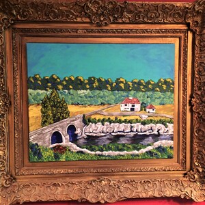 May include: An oil painting in a heavy, ornate gold-colored frame featuring a landscape with a stone bridge over a dark river, a white house with a red roof, a yellow field, and a dense green forest under a bright blue sky.