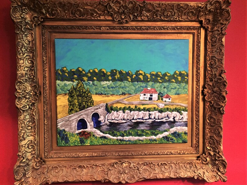 May include: An oil painting in a heavy, ornate gold-colored frame featuring a landscape with a stone bridge over a dark river, a white house with a red roof, a yellow field, and a dense green forest under a bright blue sky.