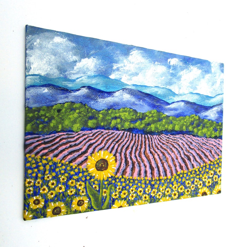 May include: A painting of a field of sunflowers in front of a lavender field. The sunflowers are yellow with brown centers and the lavender field is pink and purple. The background is a blue sky with white clouds and a mountain range in the distance.