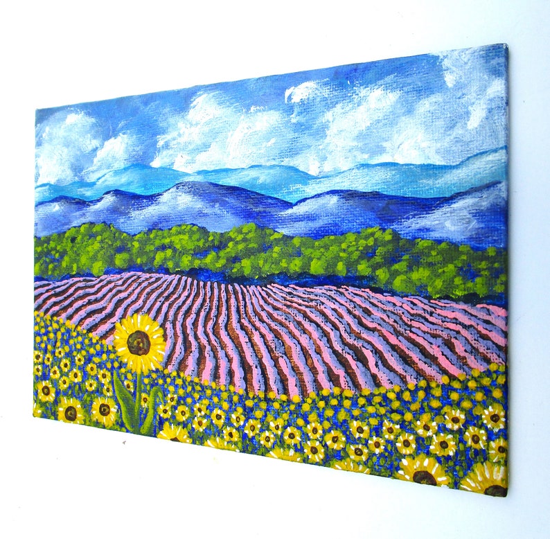 May include: A painting of a field of sunflowers in front of a lavender field. The lavender field is in rows and the sunflowers are in the foreground. There are mountains in the background and a blue sky with white clouds.