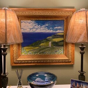 May include: Two table lamps with dark metal bases and textured tan shades flank a framed oil painting of a coastal landscape. The painting features a deep blue sea, green rolling hills, and a cloudy sky, all encased in an ornate gold frame.