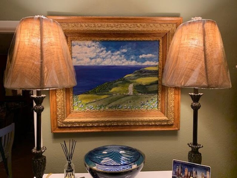 May include: Two table lamps with dark metal bases and textured tan shades flank a framed oil painting of a coastal landscape. The painting features a deep blue sea, green rolling hills, and a cloudy sky, all encased in an ornate gold frame.