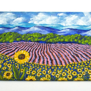 May include: A painting of a field of sunflowers in the foreground with a field of lavender in the middle ground and a mountain range in the background. The sky is blue with white clouds.