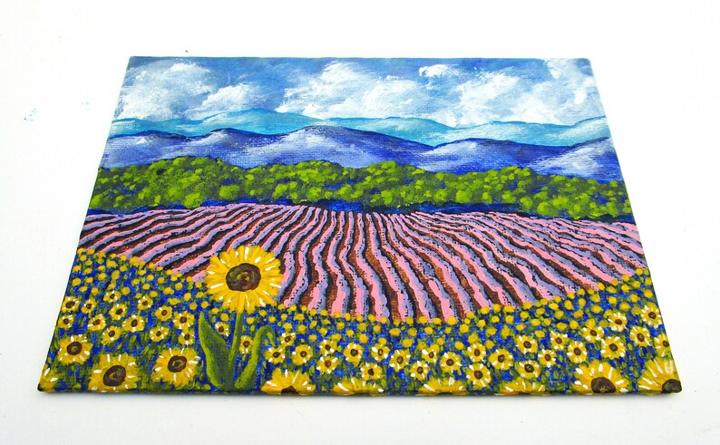 May include: A painting of a field of sunflowers in the foreground with a field of lavender in the middle ground and a mountain range in the background. The sky is blue with white clouds.