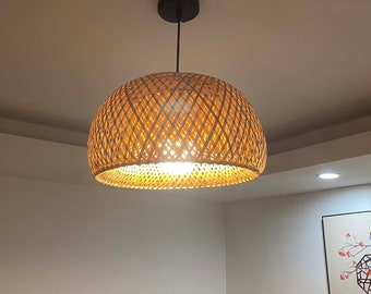 Könnte beinhalten: Eine geflochtene Bambus-Pendelleuchte mit einem natürlichen braunen Finish. Die Leuchte hängt an einem schwarzen Kabel und hat einen runden, kuppelförmigen Schirm.