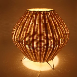 May include: A wicker lampshade with a round, bulbous shape and a woven pattern. The lampshade is lit and casts a warm glow. The lamp has a thin metal base with three legs.