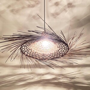 Puede incluir: Lámpara colgante ovalada hecha a mano. La pantalla es de tejido abierto, con un marco espinoso de color marrón oscuro. La lámpara está suspendida de un cable negro y emite un brillo cálido.