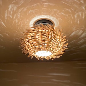 Może przedstawiać: Wisząca lampa sufitowa z naturalnych, tkanych materiałów. Lampa ma okrągły kształt z kolczastym, warstwowym wzorem. Jest zamontowana na białym suficie z ciemnym pierścieniem. Światło rzuca wzór na sufit.