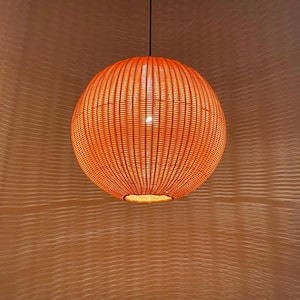 May include: Orange woven pendant light fixture. The spherical lamp is suspended from a black cord. The light is on, illuminating the woven pattern. The fixture is made of a woven material.