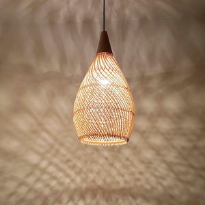 May include: A woven rattan pendant light, teardrop-shaped, with a dark brown wooden top. The light hangs from a black cord, emitting a warm glow. The shade is made of thin, light brown rattan strips, creating a textured effect.