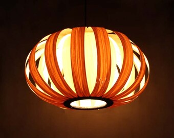 Może przedstawiać: Drewniana lampa wisząca o ciepłym, pomarańczowo-brązowym wykończeniu. Lampa wykonana jest z cienkich, zakrzywionych pasków drewna, które tworzą unikalny, geometryczny wzór. Lampa jest włączona i emituje miękkie, ciepłe światło.