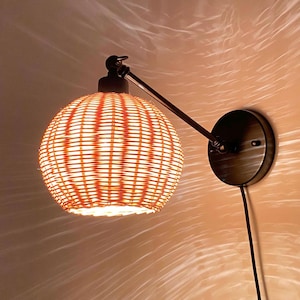 Op de afbeelding: Een wandlamp met een geweven rieten kap. De kap is lichtbruin van kleur en heeft een ronde vorm. De lamp is met een zwarte metalen arm aan de muur bevestigd.