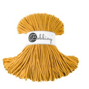 Könnte beinhalten: Eine Spule mit senfgelbem Baumwollgarn, umwickelt mit einem weißen Etikett mit dem Text "Bobbiny".