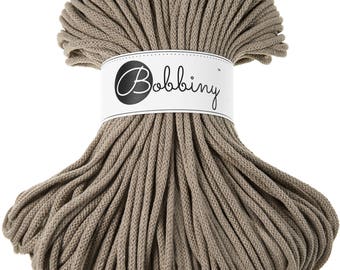 May include: A bundle of taupe-colored macrame cord. The cord is tightly wound and secured with a white label that reads "Bobbiny" in a cursive font. The cord is suitable for various crafting projects.
