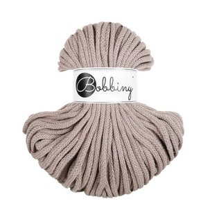 May include: A ball of taupe-colored macrame cord with a white label wrapped around it. The label features the word "Bobbiny" in a stylized script. The cord is tightly wound, and the texture is visible.