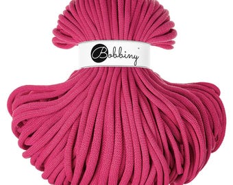 Op de afbeelding: Een felroze Bobbiny katoenen koordspoel. De spoel is gewikkeld met een wit etiket met het woord "Bobbiny" in een zwart script. Het koord is strak opgerold, waardoor een getextureerde, cilindrische vorm ontstaat, ideaal voor macramé en knutselprojecten.