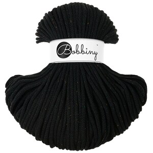 Puede incluir: Un gran carrete negro de cordón de macramé de la marca Bobbiny. El cordón está bien enrollado y tiene un aspecto ligeramente texturizado. La etiqueta es blanca con el nombre de la marca en escritura elegante. El cordón parece tener motas de color.