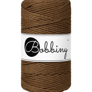 May include: A brown macrame cord spool with a white label displaying the word "Bobbiny". The cord is tightly wound, and the label is centered. The background is plain white. Ideal for crafting projects.