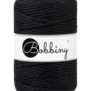 Puede incluir: Un gran carrete de hilo de macramé negro con motas de color. El hilo está enrollado sobre sí mismo y tiene una etiqueta blanca con la palabra "Bobbiny" en escritura cursiva.