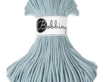 Puede incluir: Un carrete de cordón de macramé Bobbiny azul claro. El cordón está bien enrollado y sujeto con una etiqueta blanca con el logotipo de Bobbiny. El cordón está hecho de un material suave y tejido, perfecto para proyectos de manualidades.